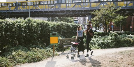 Young man carrying daughter on shoulders while woman pushing baby stroller at footpath against railway bridge