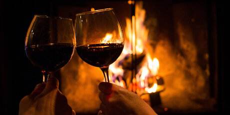 Two hands holding a full wine glass in front of a fire and clinking glasses 