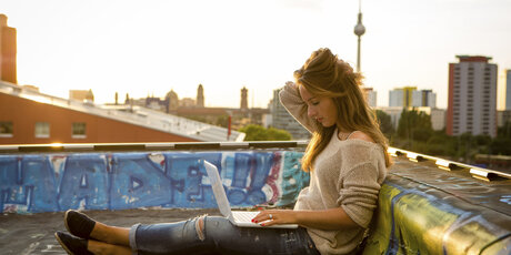 Junge Frau mit Laptop auf dem Schoß sitzt auf einem Dach