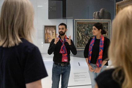 Aya und Michel tragen rot-blaue Schals des Jüdischen Museums Berlin und stehen dort in der Dauerausstellung. Sie unterhalten sich in Deutscher Gebärdensprache mit den Besuchern.