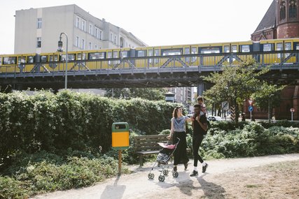 Young man carrying daughter on shoulders while woman pushing baby stroller at footpath against railway bridge