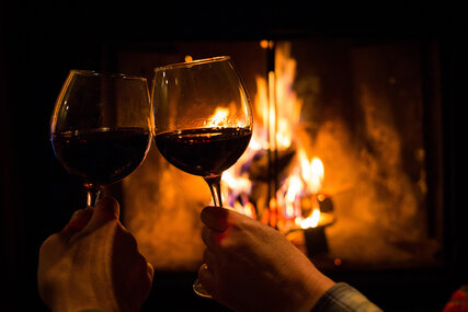 Two hands holding a full wine glass in front of a fire and clinking glasses 
