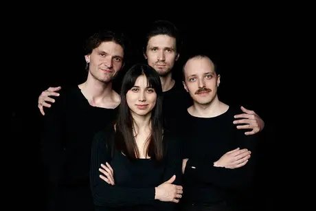 Vier schwarz gekleidete Personen umarmen sich und schauen in die Kamera