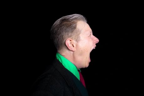 Porträt einer Person im Profil vor schwarzem Hintergrund. Der Mund ist weit aufgerissen, die Augen sind geschlossen. Kurze graublonde Haare, dazu ein schwarzer Nadelstreifenanzug über grünem Hemd und rotem Oberteil.