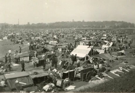Das Schwarz-Weiß-Foto zeigt eine große Ansammlung von Zelten und Wagen auf einer weitläufigen Fläche. Viele Menschen sind zu sehen, die sich in und um die Behausungen herum bewegen. Im Hintergrund ist eine Bebauung mit Bäumen und Gebäuden erkennbar. 