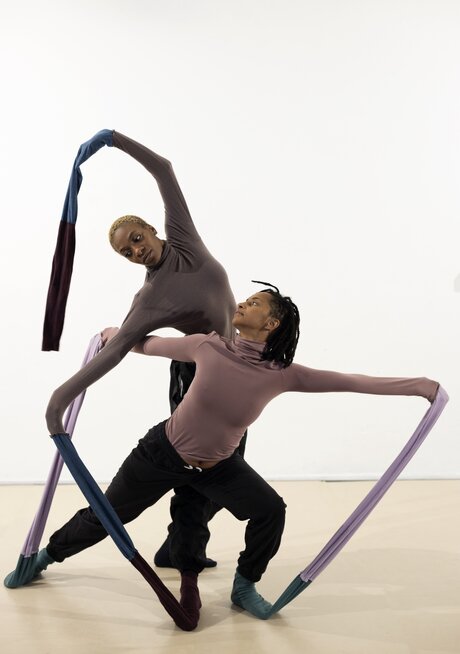 Zwei Performerinnen posieren für das Foto. Beide haben eine tänzerische Pose eingenommen. An ihren Händen und Füßen befinden sich lange Schläuche aus Stoff, die jeweils ihre eigenen Hände und Füße miteinander verbinden.