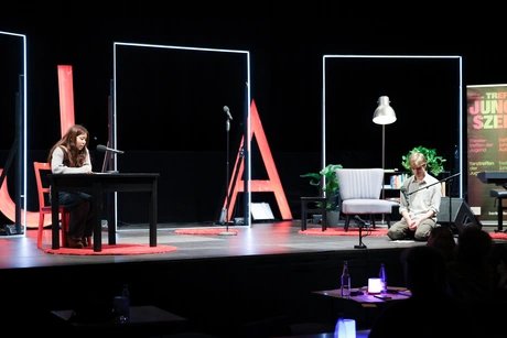 Auf der Bühne sind zwei Personen. Eine sitzt an einem Tisch vor einem Mikrofon, die andere sitzt auf den Knien vor einem Mikrofon.