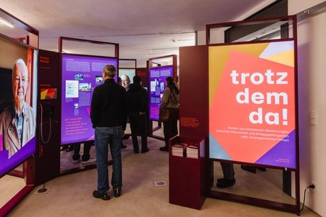 Mehrere Personen betrachten Ausstellungstafeln in einem Raum, eine Tafel zeigt den Schriftzug 'trotzdem da!' in Weiß auf rotem und gelbem Hintergrund.