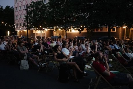Viele Menschen sitzen in Stuhlreihen auf dem Vorplatz der Berlinischen Galerie und blicken nach vorne auf eine Leinwand, die nicht auf dem Bild zu sehen ist. Es dämmert langsam.