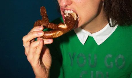 Das Bild zeigt eine junge Frau, die in eine Brezel beißt. Das obere Bildende befindet sich in der Mitte der Nase der abgebildeten Person, sodass ein Großteil des Gesichts und des Kopfes nicht sichtbar sind. Die Person trägt einen grünen Pullover, auf