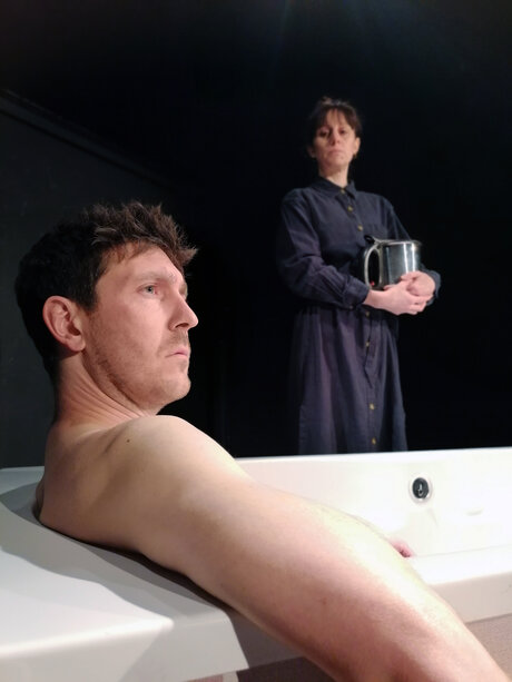 Ein Mann sitzt mit nacktem Oberkörper in einer Badewanne und hat den Mund zum Schreien aufgerissen. Eine Frau in blauem Kleid steht mit verschränkten Armen neben der Wanne und blickt auf den Mann herab.