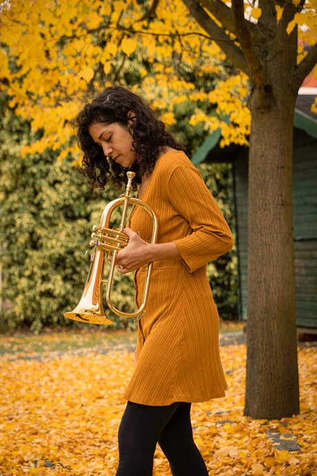 Yael Gat steht unter einem Baum in gelbem Laub. Sie hält ihre Trompete und betrachtet sie ernst.