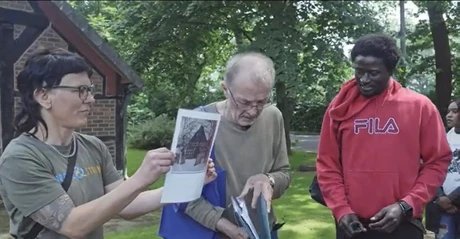 Eine Person mit tätowiertem Arm hält ein Blatt Papier mit einem Foto von einem Fachwerkhaus hoch. Daneben stehen zwei weitere Personen, eine trägt einen beigen Pullover, die andere einen roten Kapuzenpullover mit großem FILA-Schriftzug. Im Hintergrun