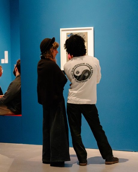 Rückenansicht von zwei Personen, die vor einer Collage von Raoul Hausmann stehen, die an einer leuchtend blauen Wand hängt.