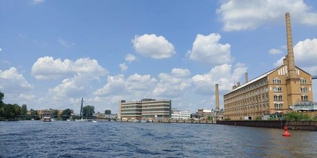 Blick auf einen Fluss mit Industriegebäuden und einem Schornstein unter blauem Himmel mit Wolken