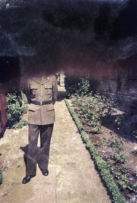 Fotografie: Farbnegativ, das eine Person in Uniform auf einem von Blumenbeeten gesäumten Weg zeigt. Der Kopf der Person und die ganze obere Hälfte der Fotografie sind dunkel angelaufen und dadurch verdeckt.