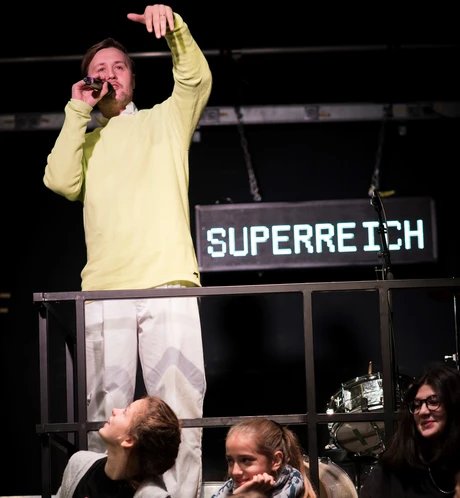 Eine Person in einem gelben Pulli und mit einer weissen Hose redet in ein Mikrophon. Hinter ihm hängt ein Schild auf dem Superreich steht.