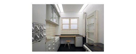 Raumfoto einer Küche mit Herd, Glastür, zugeklapptem Bügeltisch auf der rechten Seite; Schütten für unverpackte Waren aus Metall, Waschbecken und Lagerflächen auf der linken Seite. Im Hintergrund ist eine Arbeitsfläche mit Hocker, Schubladen und Fens