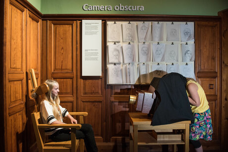 Blick in einen holzvertäfelten Ausstellungsraum. Ein Kind ist über eine große Kamera mit Holzgehäuse gebeugt, ein schwarzes Tuch über dem Kopf. Die Kamera ist auf ein Mädchen gerichtet, das in einem Lehnstuhl sitzt. An der Wand im Hintergrund hängen 