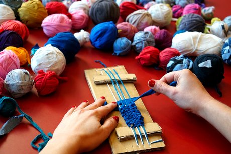 Eine Person sitzt an einem Tisch und webt an einem selbstgebauten Webrahmen aus Pappe aus einem breiten, blauen Stück Stoff ein Band. Von der Person sind nur die Hände zu sehen. Auf dem Tisch liegen viele bunte Knäule aus Stoffresten.