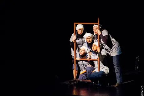 Fünf Performer*innen stehen dicht beieinander auf einer dunklen Bühne. Sie tragen Arbeitskleidung und helle Kopftücher. Eine Person steht im Vordergrund an einem Mikrofon, die anderen gruppieren sich dahinter. Gedämpftes Licht richtet sich auf die Gr