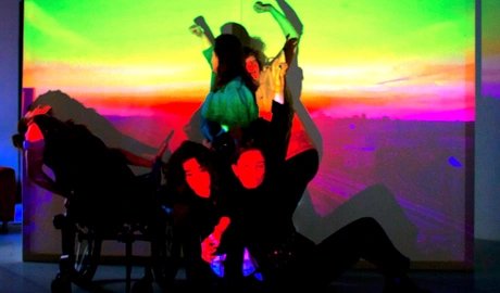 Eine Gruppe von fünf Performer:innen posiert in einer dynamischen, vielschichtigen Komposition. Sie sind vor einer Projektion, die einen leuchtenden Stadthimmel im Sonnenuntergang mit einem Farbverlauf zeigt. Die Projektion wirft starke, gesättigte F