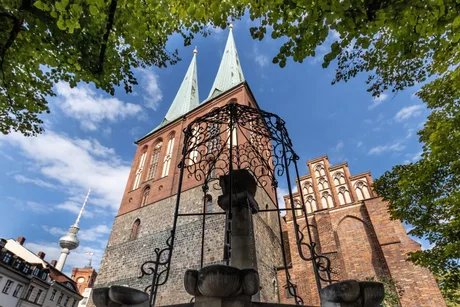 Blick durch eine schmiedeeiserne Brunnenzier auf die Türme und Giebel der Nikolaikirche, im Hinterfgrund hinter den Dächern historisierender Gebäude der Fernsehturm und der Turm des Roten Rathauses