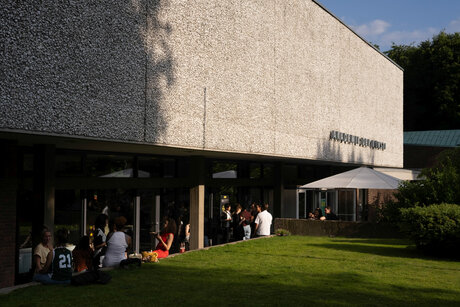 Außenansicht des Gebäudes der Akademie der Künste im Hanseatenweg. Einige Besucher*innen stehen vor dem Eingangsbereich und unterhalten sich. Die Sonne scheint, das Gras der Wiese auf dem Vorplatz leuchtet grün.