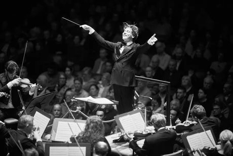 Dirigent Klaus Mäkelä breitet die Arme vor dem Orchester aus, die Musiker:innen streichen ihre Geigen und im Hintergrund sieht man das Publikum.