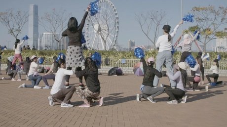 Video-Still: Eine Gruppe von Menschen im Freien mit blauen Pompons, einige kniend und andere stehend, vor einem Riesenrad und Hochhäusern in einer Stadtlandschaft.