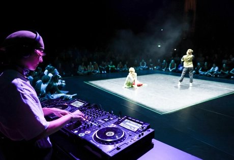 Menschen tanzen auf einer beleuchteten Bühne in einem sonst dunklen im Raum. Im Vordergrund ist angeschnitten ein*e DJ an Turntables zu sehen.