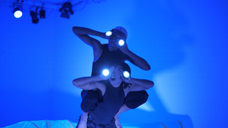 Der Raum ist in blaues Licht getaucht. Zwei Performerinnen halten sich die Hände vor die Augen. Auf ihren Handrücken befinden sich runde, leuchtende Kreise. Die eine Performerin sitzt aufrecht auf dem Rücken der anderen.