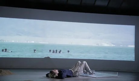 Zu sehen ist eine Person, die vor einer Videoprojektionsleinwand auf dem Boden liegt. Es wird eine Aufnahme im Ultabreitformat von in einem Gewässer badenden Menschen projiziert. Die Person, die auf dem Boden liegt, hat ihre Beine aufgestellt und ihr