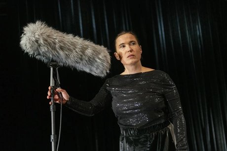 Frau in glitzerndem schwarzen Outfit hält ein großes, mit Fell verkleidetes Mikrofon und schaut konzentriert nach vorn.