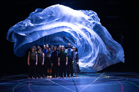 Eine Gruppe von Tänzern steht in einem schwarzen Raum unter einer blau beleuchteten Schwebeplastik, die über ihren Köpfen schwebt