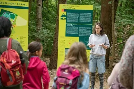 Eine Frau erzählt vor einem Schild stehend die Geschichte der mittelalterlichen Siedlung im Museumsdorf Düppel