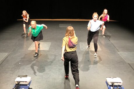 Fünf junge Frauen in sportlicher Kleidung vollziehen Laufbewegungen auf einer Bühne.