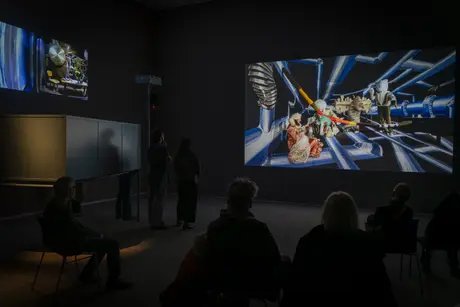 Foto: Ein paar Menschen sitzen und stehen in einem Videoraum und schauen auf eine große Leinwand.