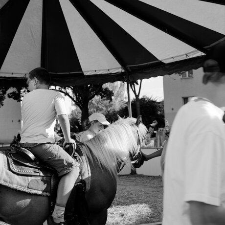 Schwarz-Weiß-Fotografie: Seitenansicht von einem Jungen auf einem Pferd unter einem gestreiften Baldachin. Im Hintergrund sind weitere Menschen zu sehen.
