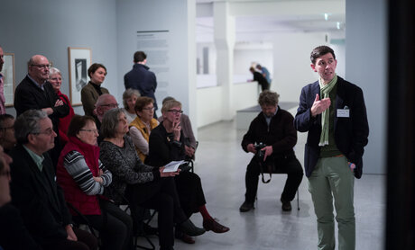 Viele Menschen stehen und sitzen im Ausstellungsraum und schauen auf eine Person, die spricht und auf ein Gemälde deutet. Zirkeltraining in der Berlinischen Galerie