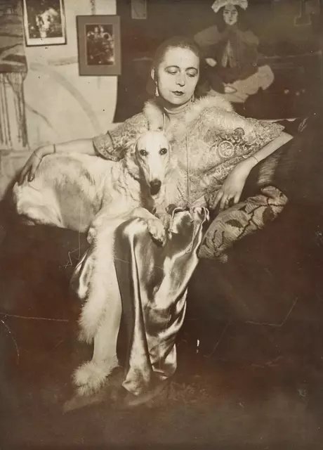 Selbstportrait der Künstlerin Marta-Astfalck-Vietz als Schwarz-Weiß-Fotografie. Sie sitzt in einem glänzenden Kleid mit Pelzbesatz auf einem Sofa. Auf ihrem Schoß sitzt ein Hund mit hellem Fell. Im Hintergrund sind Gemälde und dekorative Elemente zu 