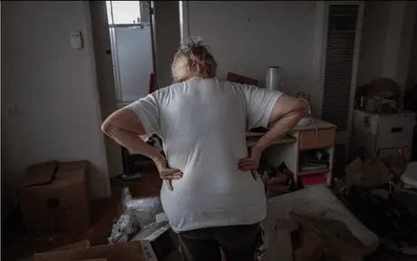 Das Foto zeigt eine Frau, die mit dem Rücken zur Kamera in einem unordentlichen, chaotischen Raum steht. Sie trägt ein weißes T-Shirt und dunkle Hosen und stützt ihre Hände in den unteren Rücken, als sei sie erschöpft oder habe Schmerzen.