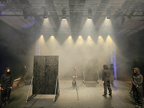 Tänzer:innen in schwarzer Kleidung stehen auf einer nebligen Bühne. Einige blicken nach vorne, andere sind von hinten zu sehen. Zudem befinden sich zwei schwarze Wandaufsteller auf der Bühne.