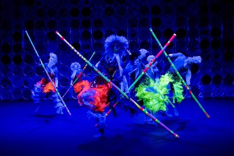 Die rüschigen Kostüme und langen Stäbe von den Tänzer:innen leuchten in bunten Neonfarben.