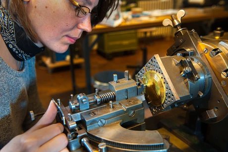 Eine Frau bedient eine Guillochier-Maschine. Mit der rechten Hand bewegt sie die Graviernadel der Maschine über das eingespannte Metallstück.