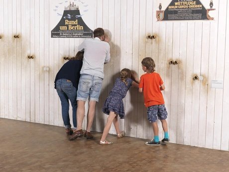 Eine vierköpfige Familie steht vor einer weißen Wand. Durch Gucklöcher in der Wand werfen sie einen Blick auf dahinter angebrachte Fotografien historischer Flugplätze. Auf der Wand sind historische Werbeplakate für Wettflüge aufgehängt.