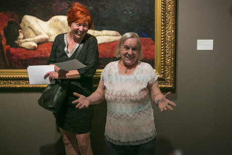 Zwei Frauen stehen vor einem Gemälde. Sie lachen in die Kamera. Eine Frau macht eine einladende und offene Geste.