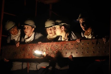 Fünf Performer*innen tragen Schutzhelme und Arbeitskleidung. Sie lehnen sich gemeinsam über eine schwere Metallplatte mit Nieten. Eine kleine Lichtquelle beleuchtet ihre Gesichter von unten.
