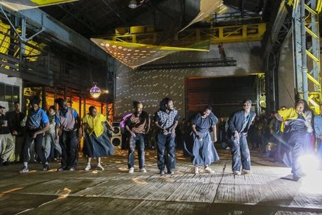 Eine Gruppe von Menschen tanzt energisch in einem großen Veranstaltungsort im Industriestil, der von bunten Lichtern und Discokugeln beleuchtet wird, mit Zuschauern im Hintergrund.