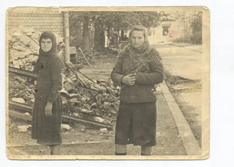 Das Foto zeigt zwei junge Mädchen vor dem Hintergrund von Trümmern. Das Mädchen links trägt ein dunkles Kleid und ein Kopftuch. Das Mädchen rechts trägt ein dunkleres Kleid, Strümpfe und einen gehäkelten Schal. Der Hintergrund deutet auf Zerstörung u